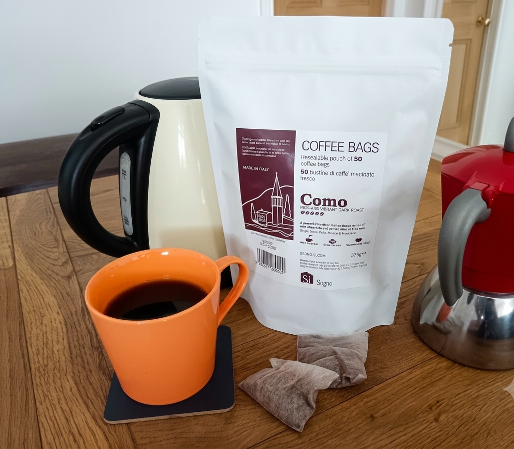 a cup of Como coffee bags next to a kettle and a moka pot
